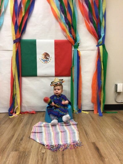 Cathy's grandson sitting on a blanket on the floor in front of the Mexican flag, age 6 months.