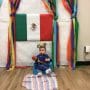 A baby sitting on a blanket on the floor in front of the Mexican flag