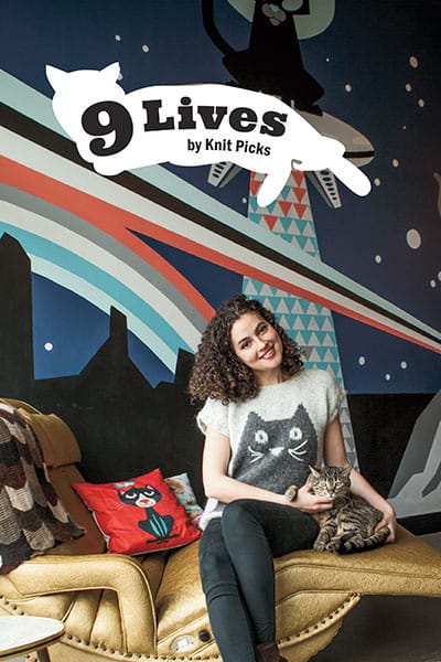 the cover of 9 Lives by knit picks: a model sits with a cat. The model is wearing a hand-knitted sweater with a cat motif on frong.