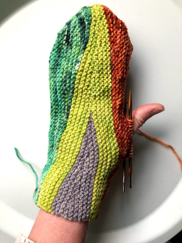 One of Lee's Unfinished Objects: A hand wearing a partially-knitted mitten in green, chartreuse, gray, and red