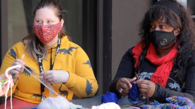 Two knitters sit next to each other outdoors and work on knitting projects.