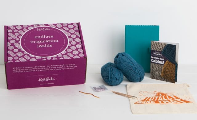 The contents of a boxed knitting kit including 2 balls of blue wool yarn, a Learn to Knit Cables! book, needle tips cable needle, teal chart keeper, and canvas project bag.