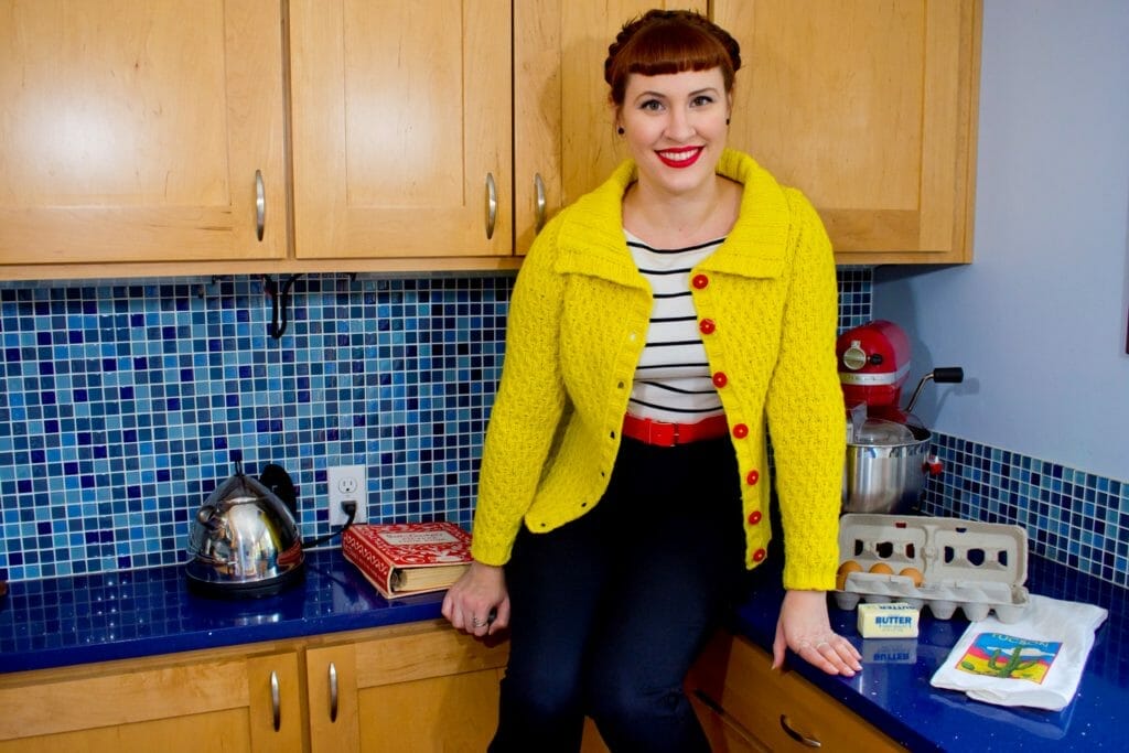 Andi Satterlund sit on the kitchen counter in a blue kitchen. She is wearing a bright yellow cardigan and has bright red lipstick, dark hair, and 1950s-style short bangs.