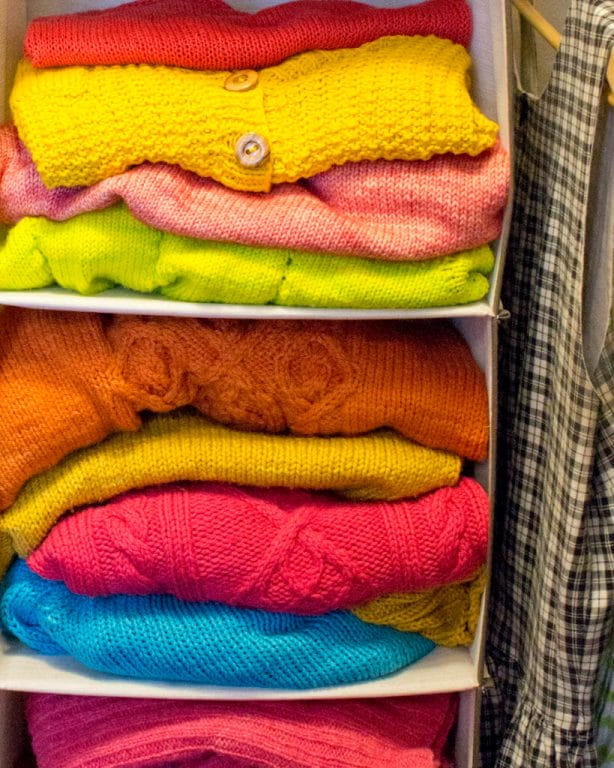 A hanging wrack in a closet is stuffed full of folded hand-knit sweaters in bright pinks, warm yellows, orange, and blue.