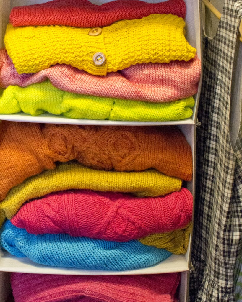 A hanging wrack in a closet is stuffed full of folded hand-knit sweaters in bright pinks, warm yellows, orange, and blue.