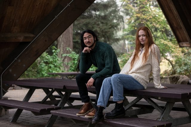 A man and woman in matching cabled sweaters sit on a picnic table.