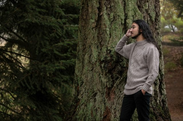 A man leans against a tree, wearing a cabled pullover sweater.