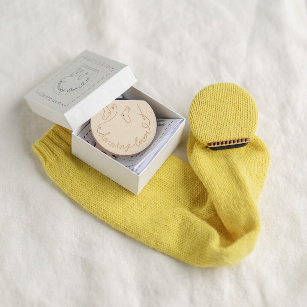 an unassembled darning loom in the box is next to a set up darning loom with a yellow sock stretched over it.