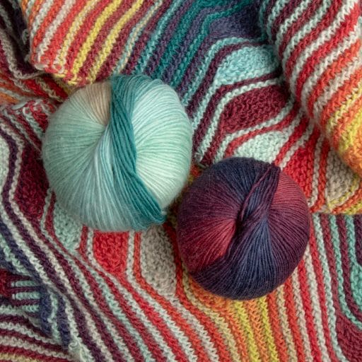 Sobre la envoltura se colocan ovillos de lana Chroma en las combinaciones de colores Seahorse (tonos de azul y amarillo) y Sedona (tonos de rojo, naranja y morado). 