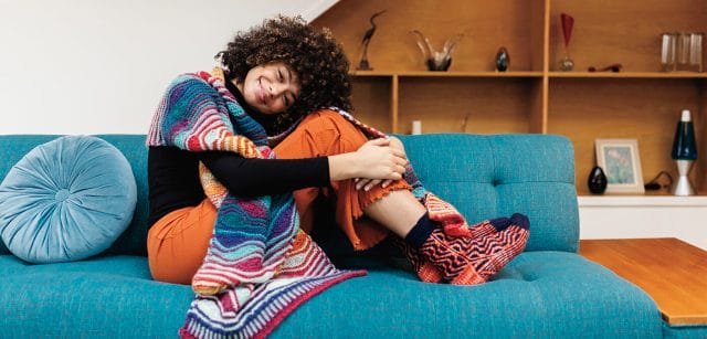 A Black woman with an afro wears a muiltcolor handknit wrap while sitting on a couch in a 1970s house. 