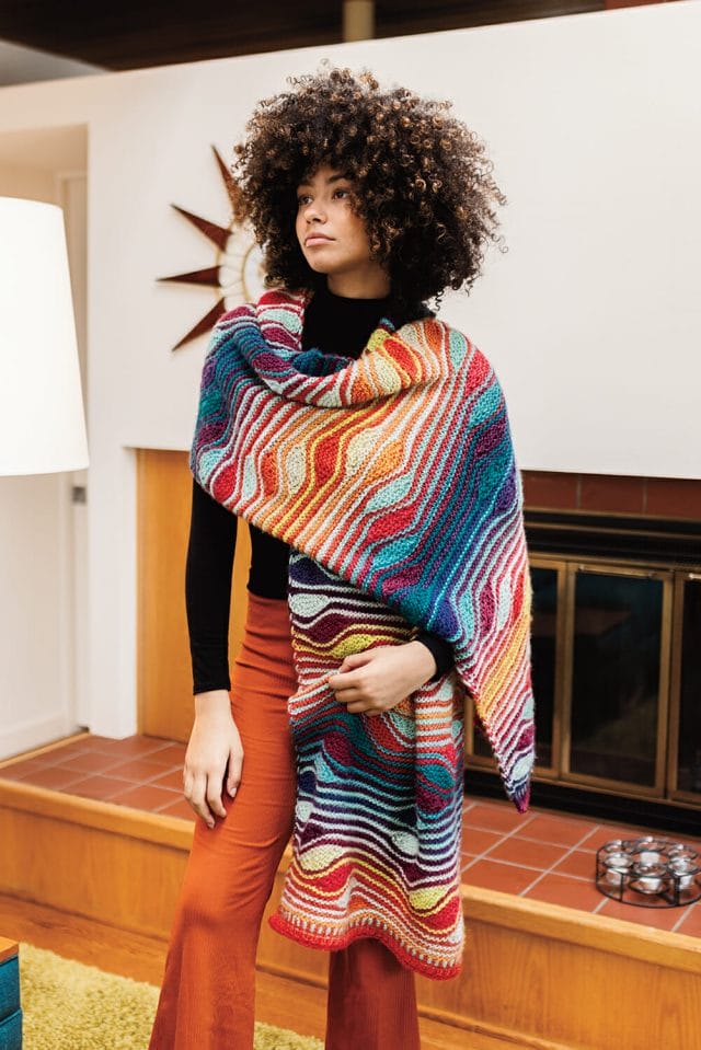Una mujer negra con afro viste un abrigo multicolor en una casa de los años 70. 
