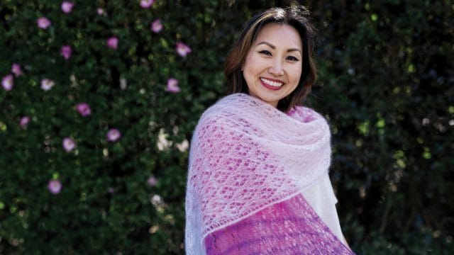 An Asian-American woman smiles while wearing a lace wrap knit with 4 shades of pink mohair yarn. 