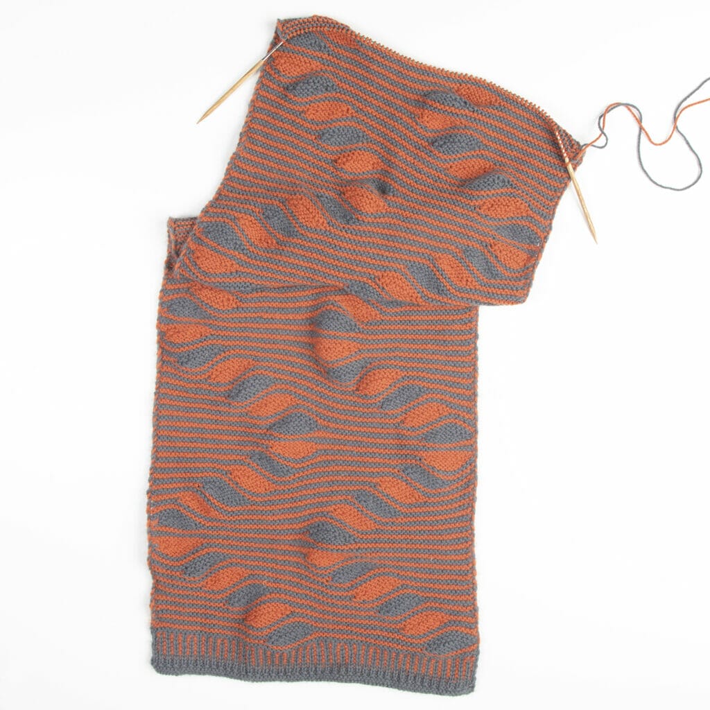An incomplete handknit wrap on knitting needles with squiggly stripes in orange and gray.