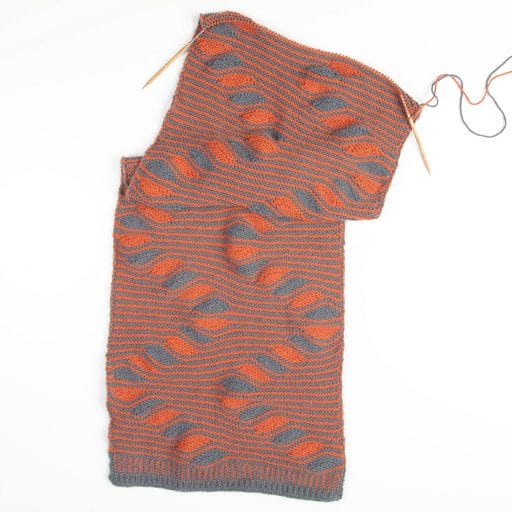An incomplete handknit wrap on knitting needles with squiggly stripes in orange and gray.