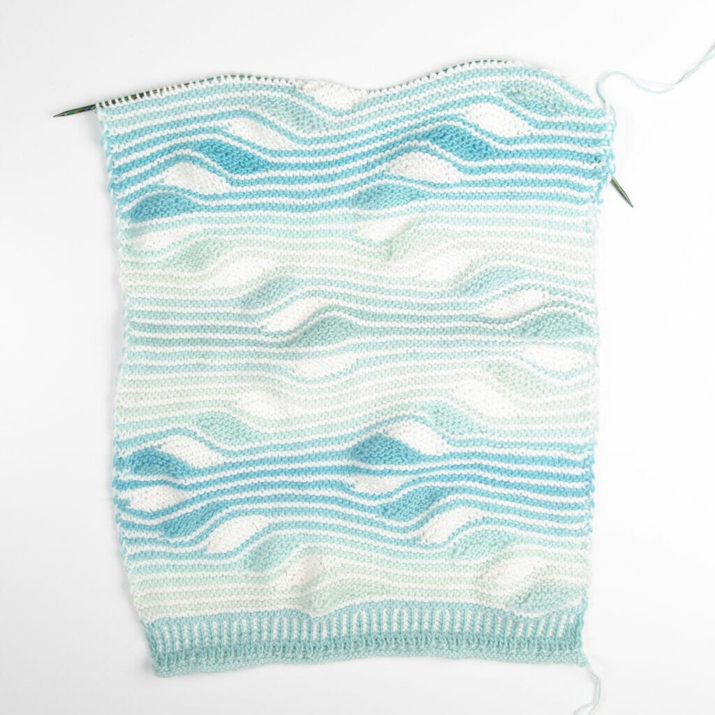 An incomplete handknit wrap on knitting needles with squiggly stripes in shades of blue and solid white. 