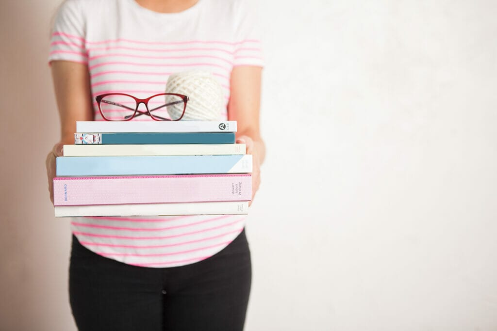 A woman holds a stack of books with glasses and a yarn ball balanced on top.