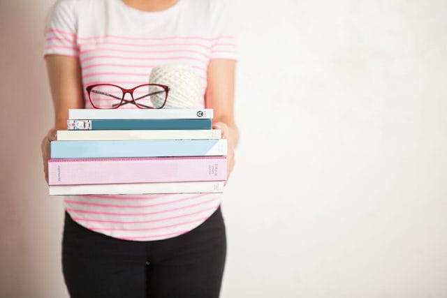 A woman holds a stack of books with glasses and a yarn ball balanced on top. 